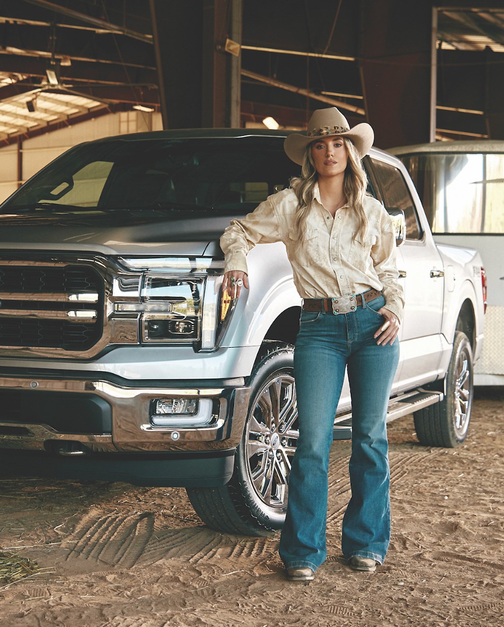 Artist in a cowboy hat standing beside a silver Ford F-150 inside a rodeo barn.