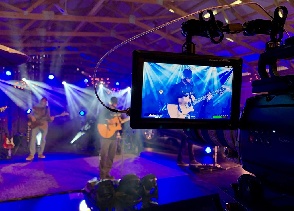Behind the scenes of a Ford Music livestream concert: a camera monitor frames an artist playing acoustic guitar under blue stage lights.